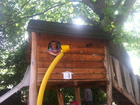 Ein Holzspielehaus auf Stelzen mit Rutsche. Aus einem Fenster kippt ein Junge etwas aus einem gelben Eimer in ein langes gelbes Rohr.
