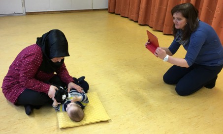 Eine Frau filmt mit einem iPad, wie eine andere Frau mit Kopftuch mit ihrem auf dem Boden liegenden Baby spielt.