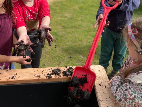 Das Hochbeet wird mit einer roten Schaufel befüllt. Kinder zeigen matschige Hände.