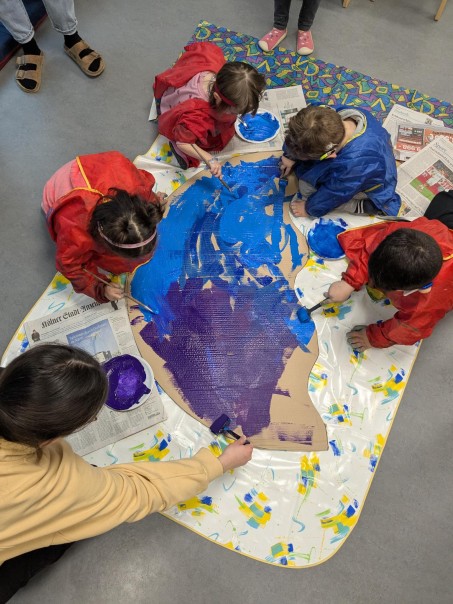 Eine große Pappe wird von Kindern und Erwachsenen blau und lila angemalt.