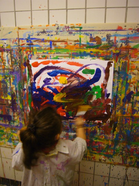 Ein Kind malt mit vielen bunten Farben auf ein großes Papier, das an der Wand befestigt ist.