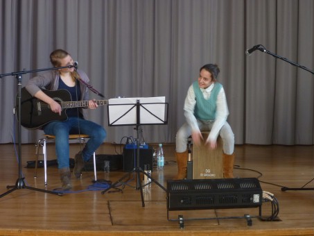Eine Schülerin spielt auf dem Cajon. Sie sitzt auf dem Cajon und Laura Müller sitzt mit ihrer Gitarre links von ihr.