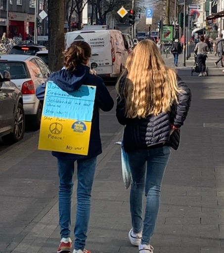 Zwei Mädchen von hinten, die mit Plakat und Spendendose auf dem Gehweg gehen.