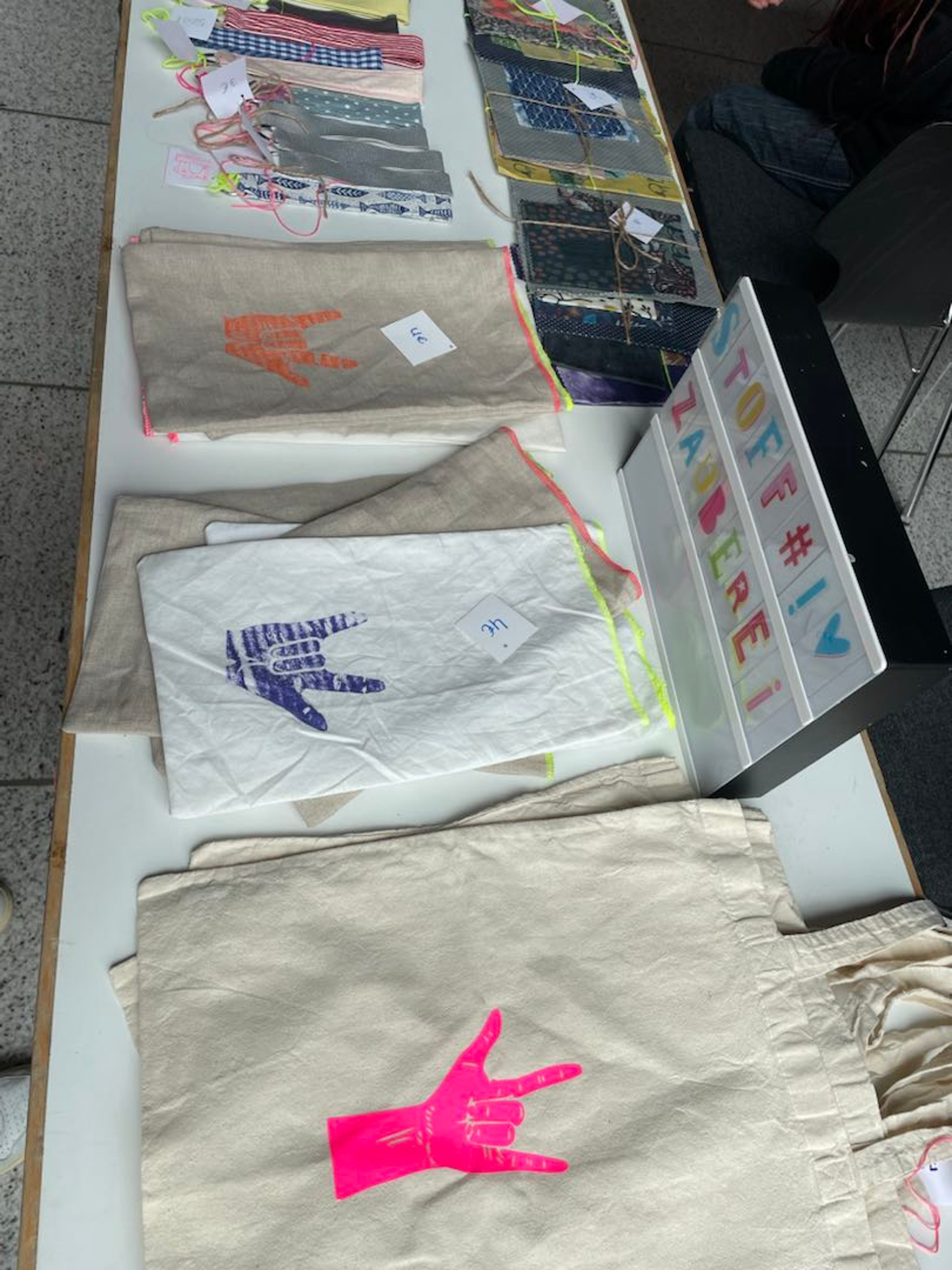 Auf einem Tisch liegen Stoffbeutel und Taschen, die mit der ILY-Hand bedruckt wurden.