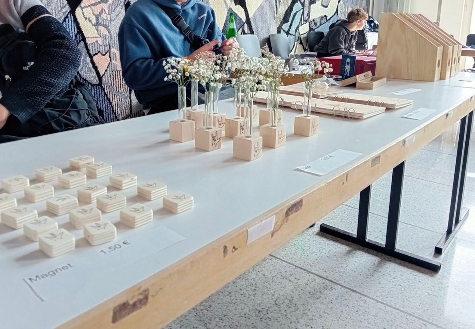 Auf einem Tisch liegen kleine, quaderförmige Holzmagnete, in die die ILY-Hand eingebrannt wurde und Vasen aus Holzklötzen mit Reagenzgläsern.