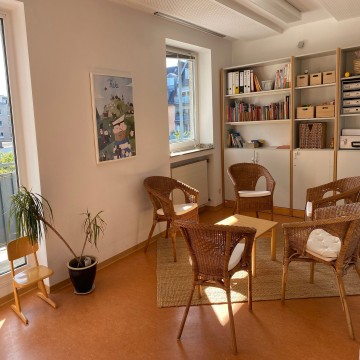 Blick in den Beratungsraum der Beratungsstelle: ein kleiner Tisch mit fünf Korbsesseln. Hinten an der Wand ein Regal mit Büchern und Schubkästen.