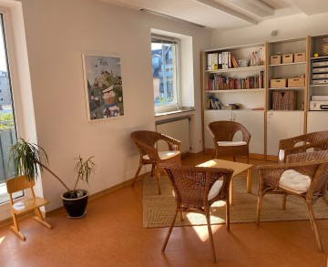 Blick in den Beratungsraum der Beratungsstelle: ein kleiner Tisch mit fünf Korbsesseln. Hinten an der Wand ein Regal mit Büchern und Schubkästen.