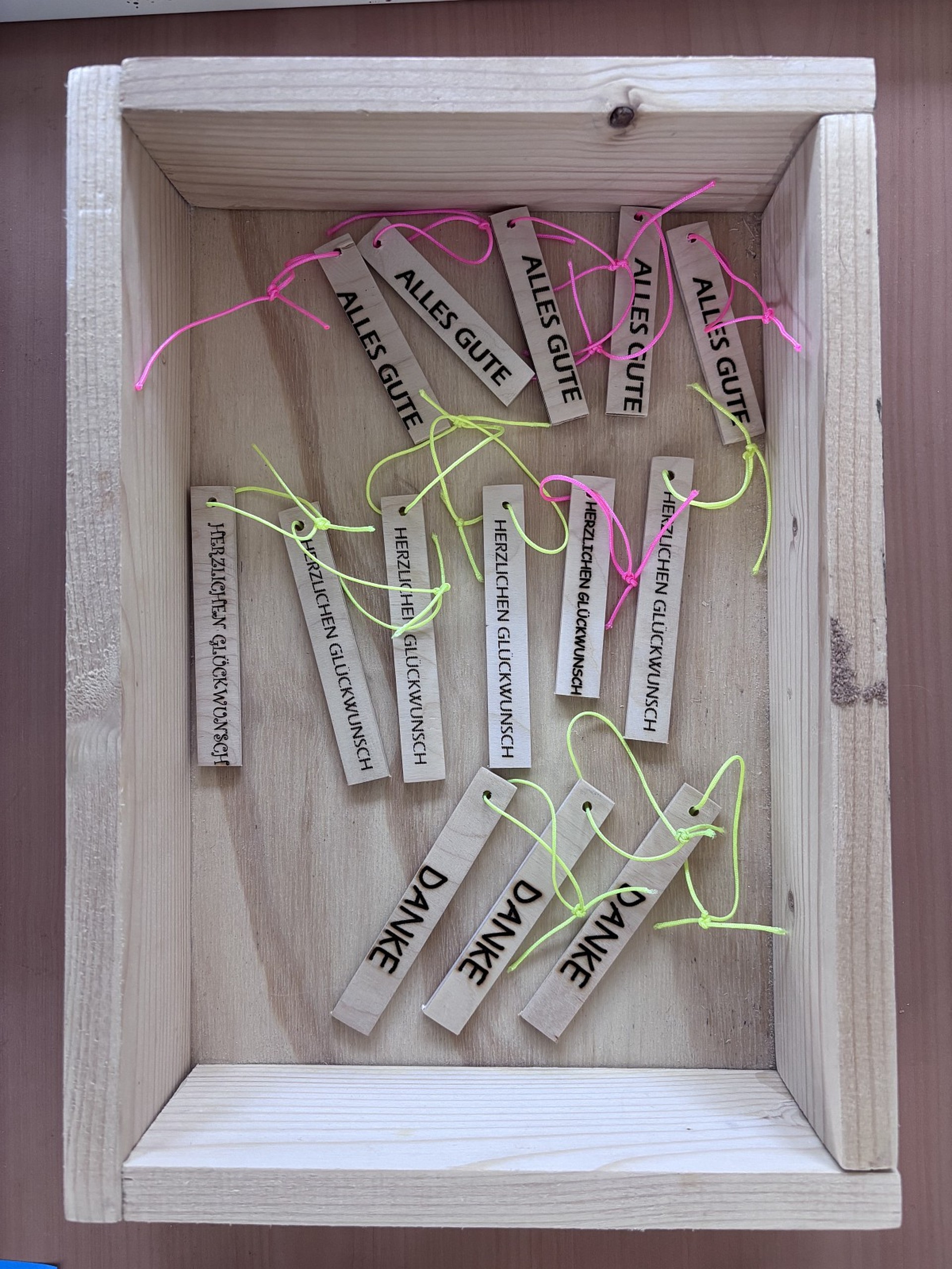 In einer Holzkiste liegen kleine Anhänger aus Holzbrettchen auf denen "Alles Gute", "Herzlichen Glückwunsch" oder "Danke" steht.