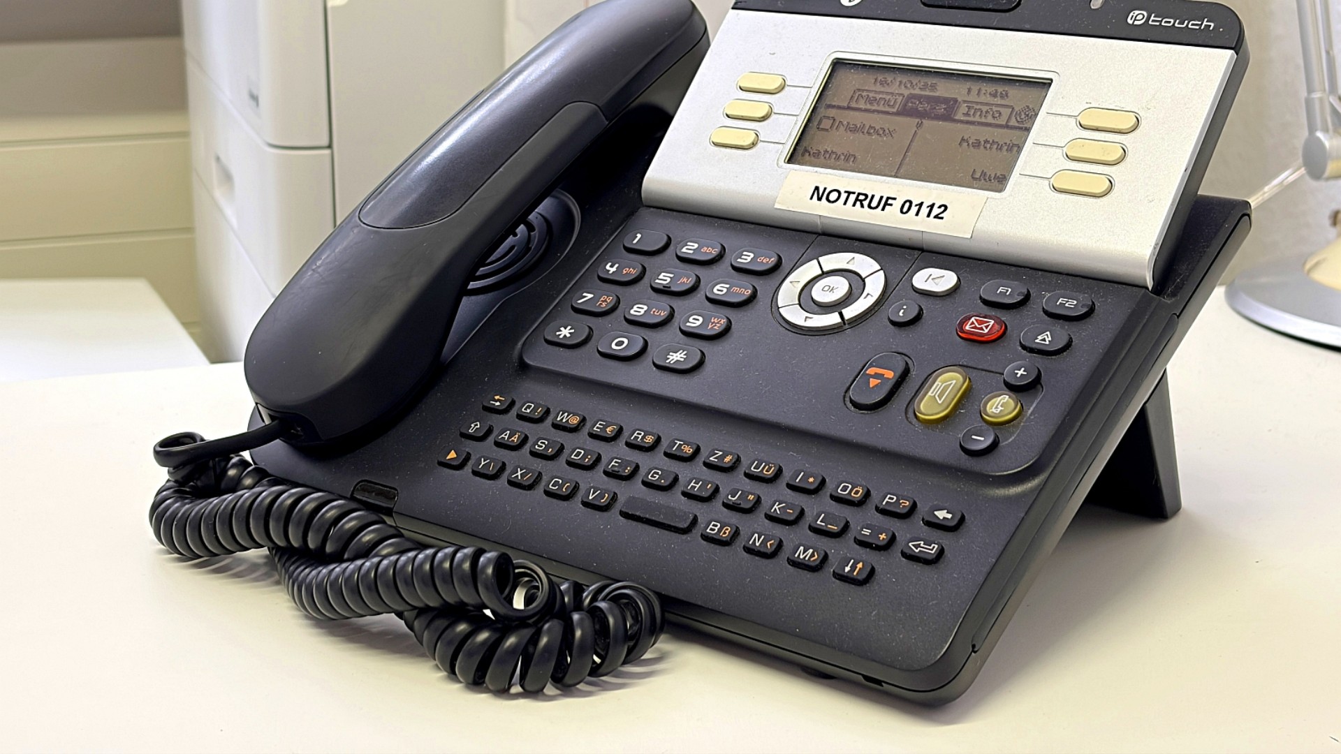 Ein schwarzes Telefon mit vielen Tasten. Im Hintergrund ein Kopierer.