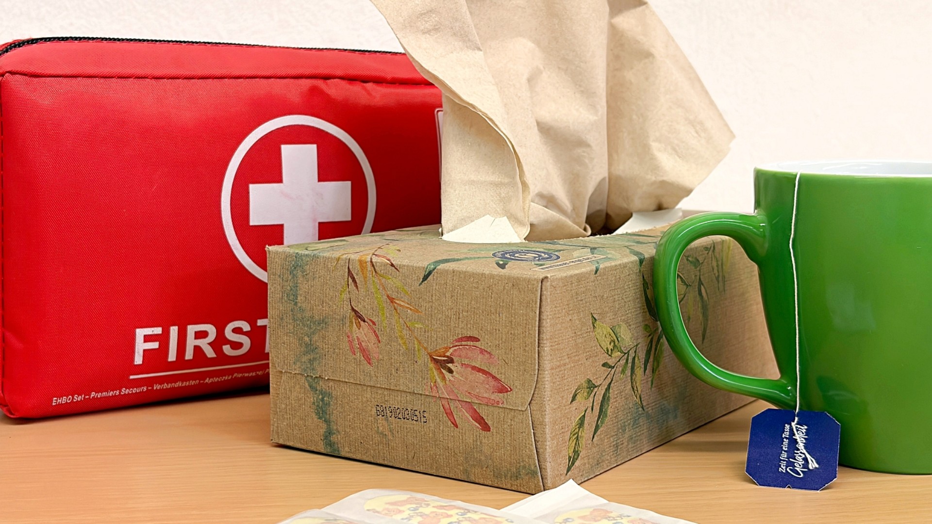 Auf einem Tisch stehen eine rote Erste-Hilfe-Tasche, eine Box mit Taschentüchern, eine grüne Tasse mit Tee-Beutel und ein paar Pflaster