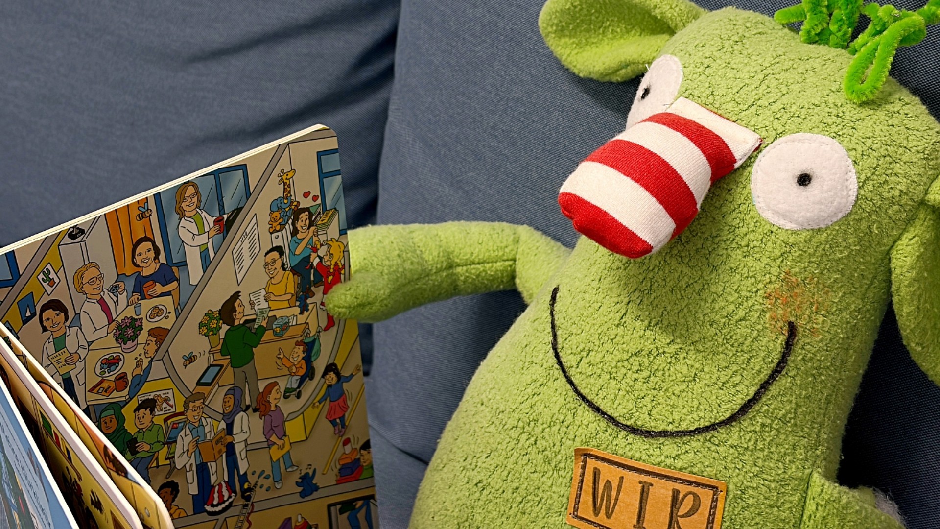 Ein grünes Kuscheltier mit Namensschild "Wir" sitzt auf einer blauen Couch und haöt ein Bilderbuch in der Hand