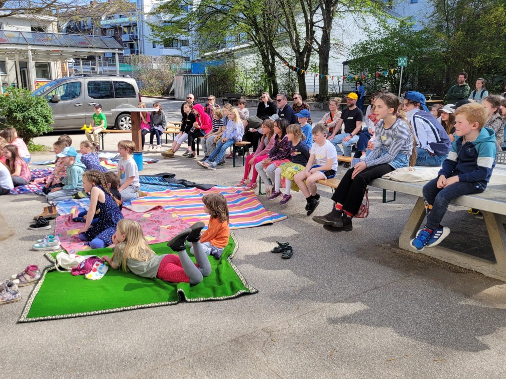 Etwa 50 sommerlich gekleidete Kinder und Eltern sitzen auf Decken, Bänken und einer Tischtennisplatte und scheun interessiert auf etwas, das nicht im Bild zu sehen ist
