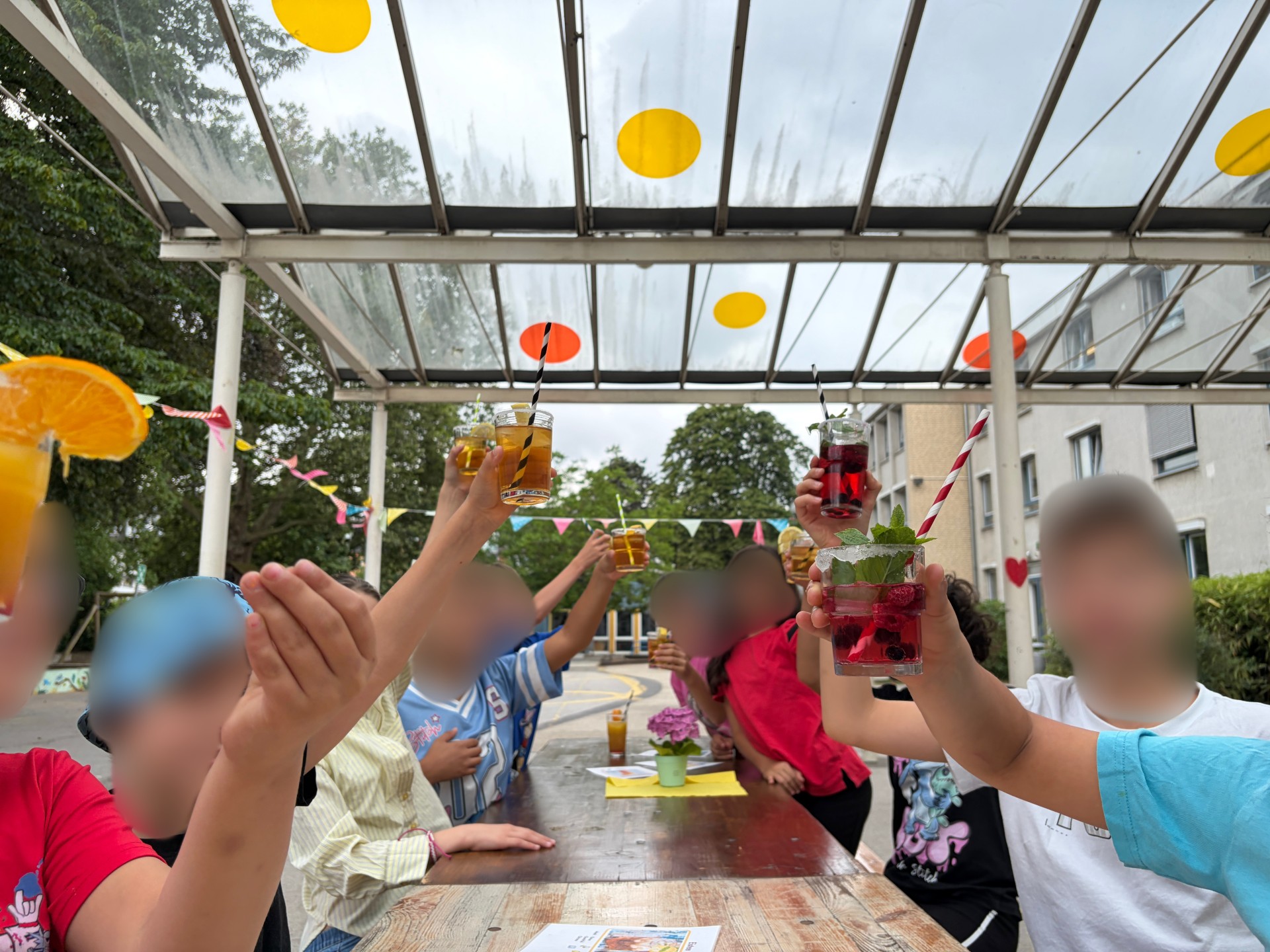 Foto auf Kopfhöhe einer Klasse, die an Bierzelt-Garnitur sitzt. Alle halten ihren bestellten Cocktail in die Höhe. Die Stimmung ist heiter und bunt.