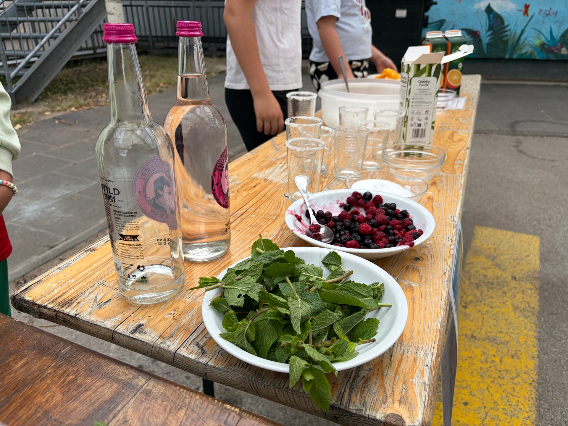 Foto von Schrägansicht auf Bierbank-Tisch. Zentral zu sehen ist Cocktail-Bar mit vorbereiteten Tellern für frische Minze und Beeren, Gläsern und Mixgetränken.
