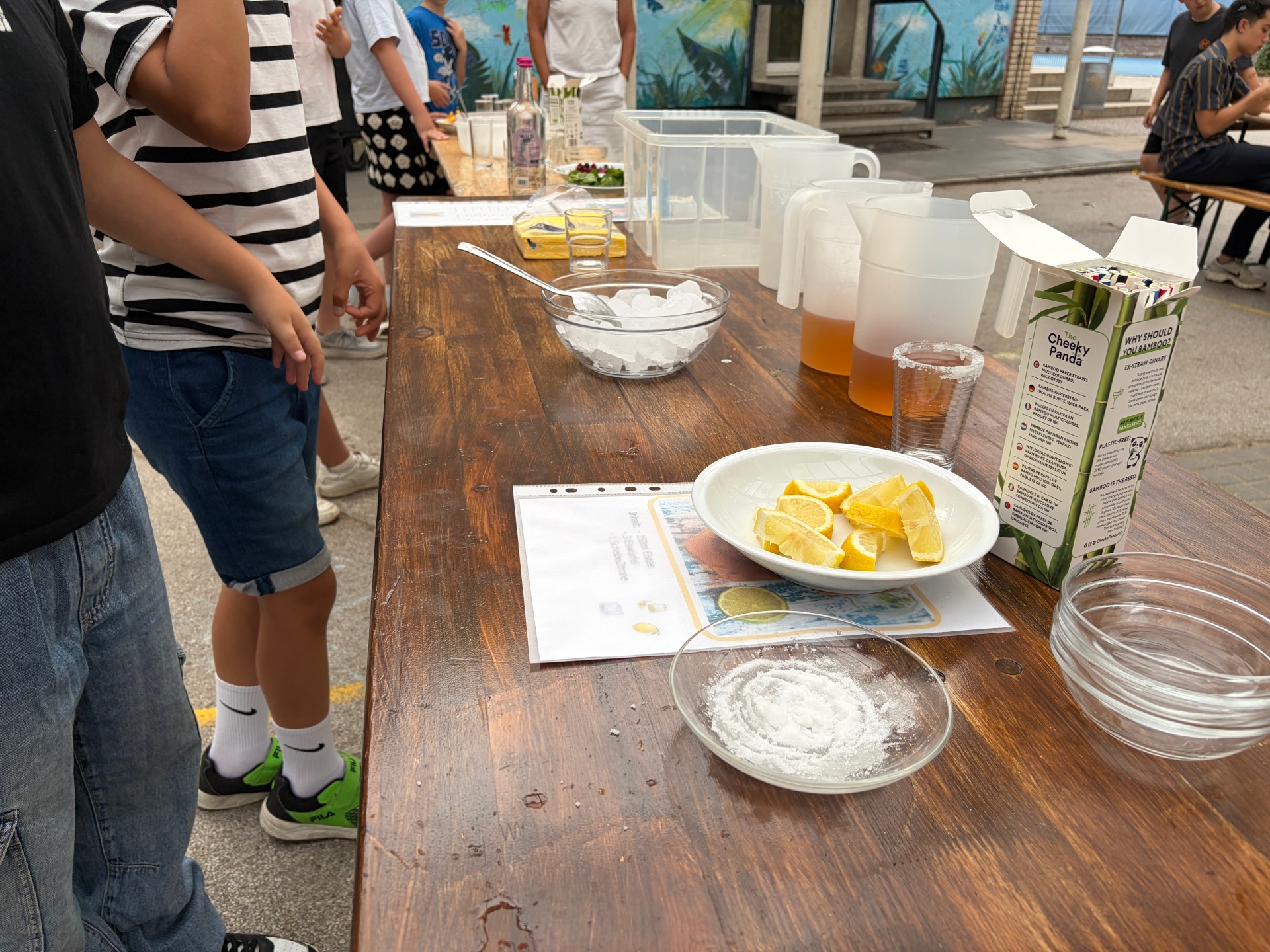 Foto von Schrägansicht auf Bierbank-Tisch. Zentral zu sehen ist Cocktail-Bar mit vorbereiteten Tellern für frische Zitronen und Mischbechern für den Eistee.
