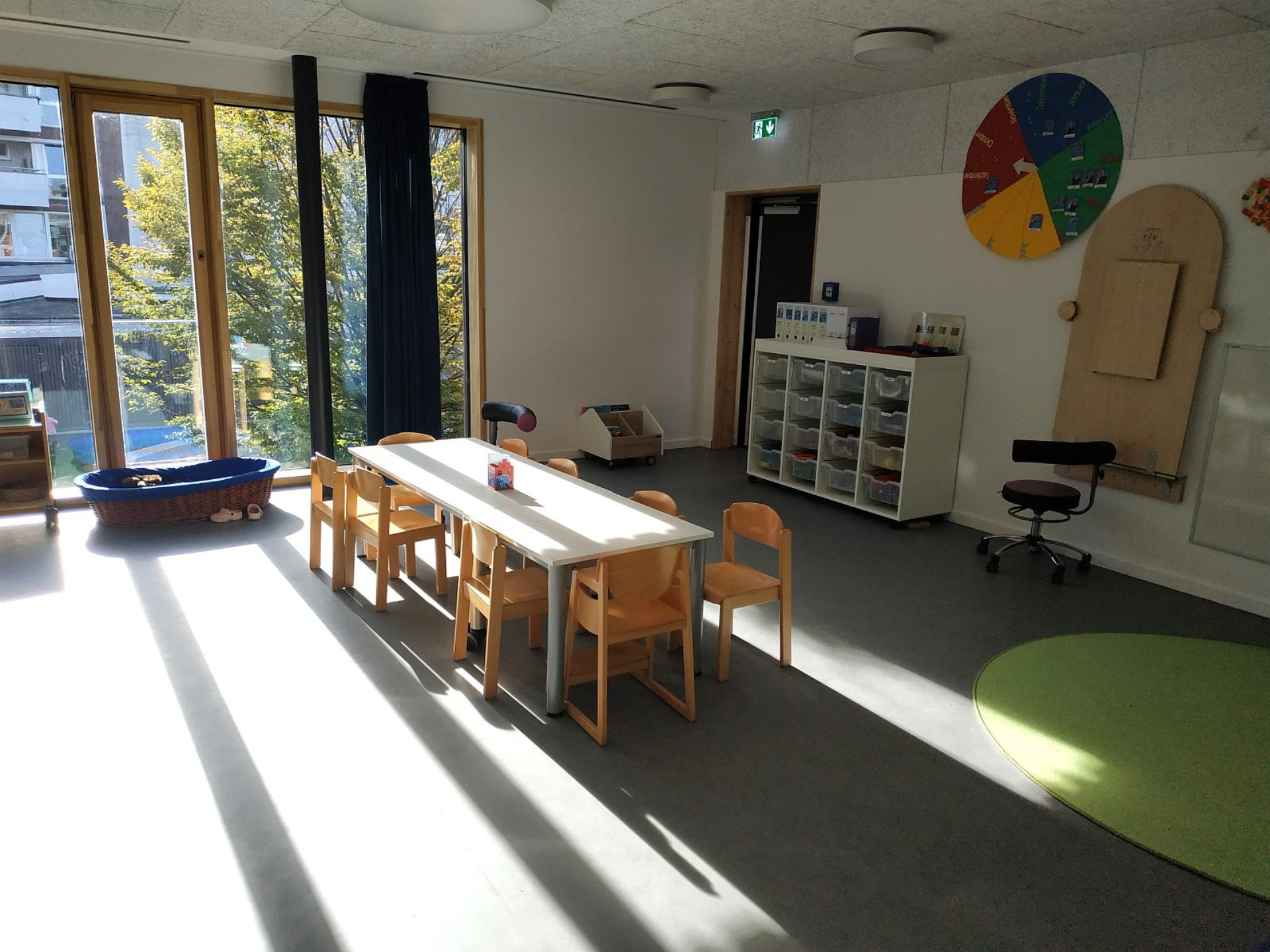 Lichtdurchfluteter Gruppenraum des Kindergartens: Gruppentisch mit Stühlen, Regal, ein runder Teppich. An der Wand hängt eine große Jahreszeitenuhr.