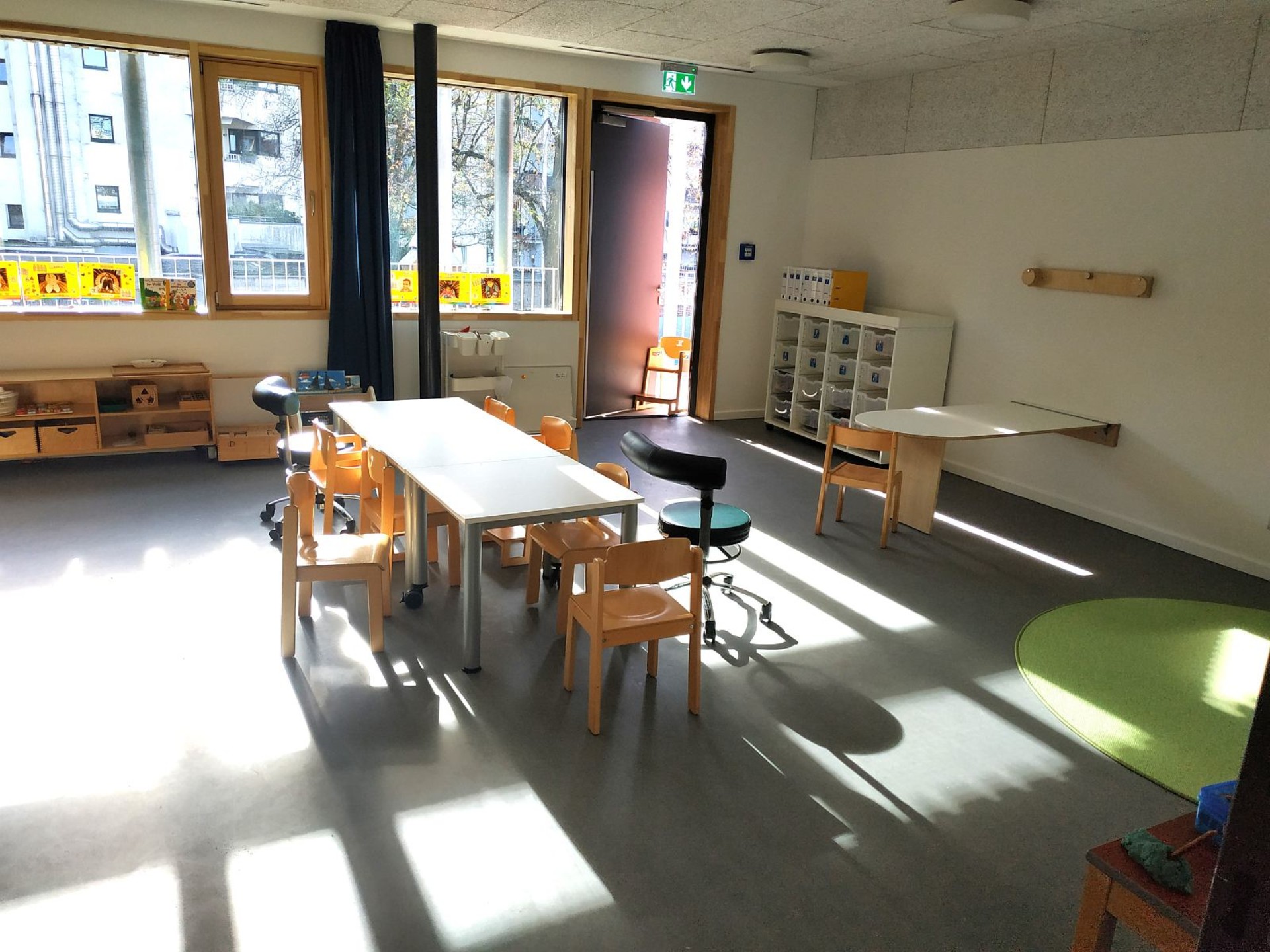 Heller Raum des Kindergartens mit Tischen und Stühlen, gelben Bildern an den Fenstern und Regale mit Spiel und Bastelmaterial.