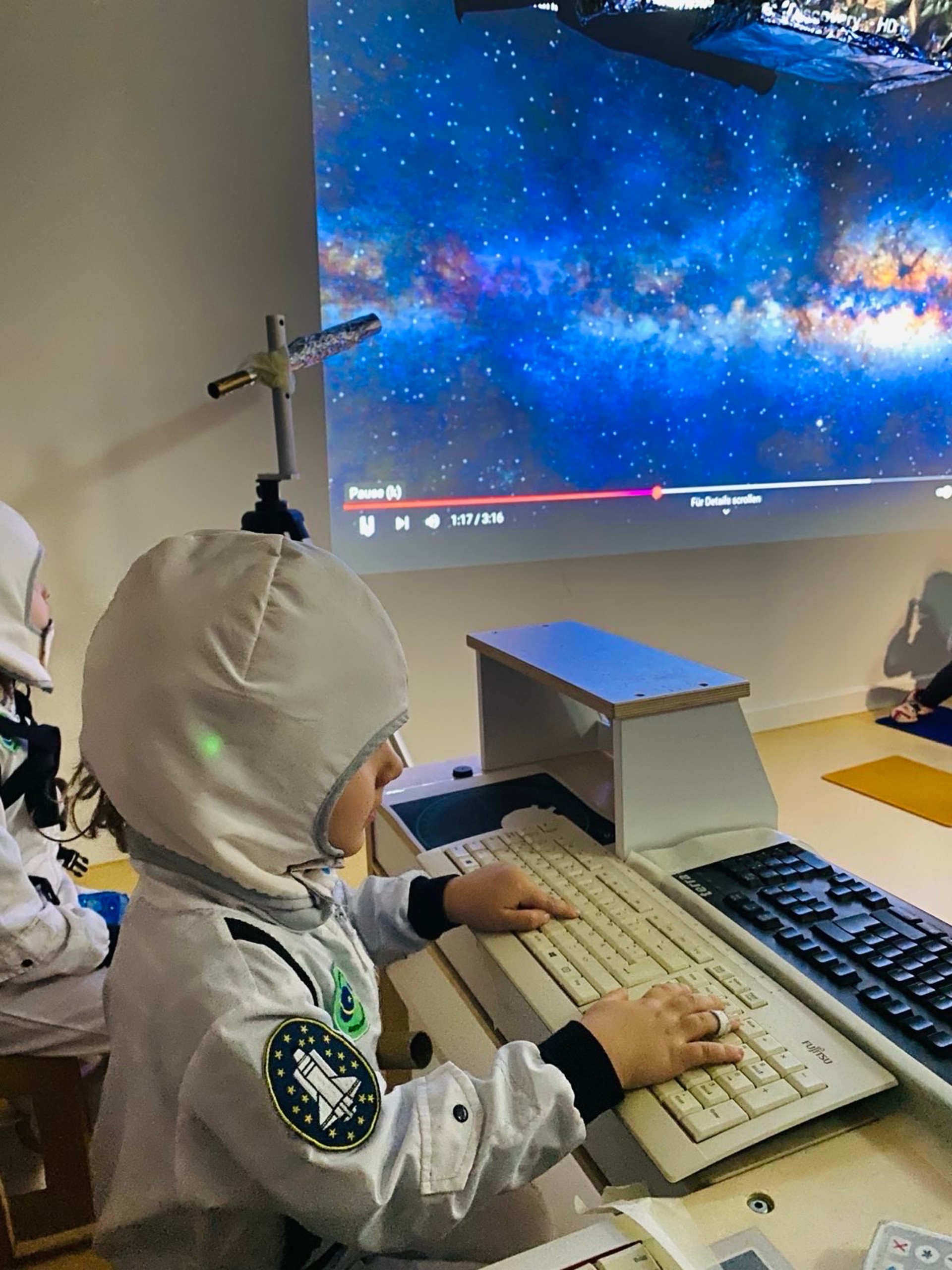 Kind in Astronautenkleidung sitzt in einer aus Tastaturen gebauten Kommandozentrale. An der Wand ist eine Projektion des blauen Weltalls zu sehen.