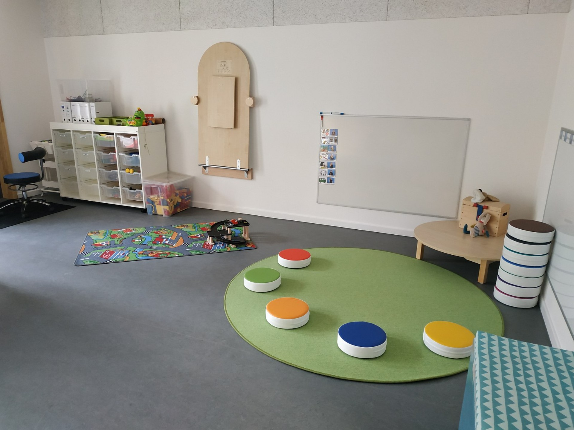 Blick in einen Gruppenraum des Kindergartens: Ein grüner runder Teppich mit bunten Sitzkissen, ein Spieleteppich und ein Regal mit Spiel- und Bastelmaterial.