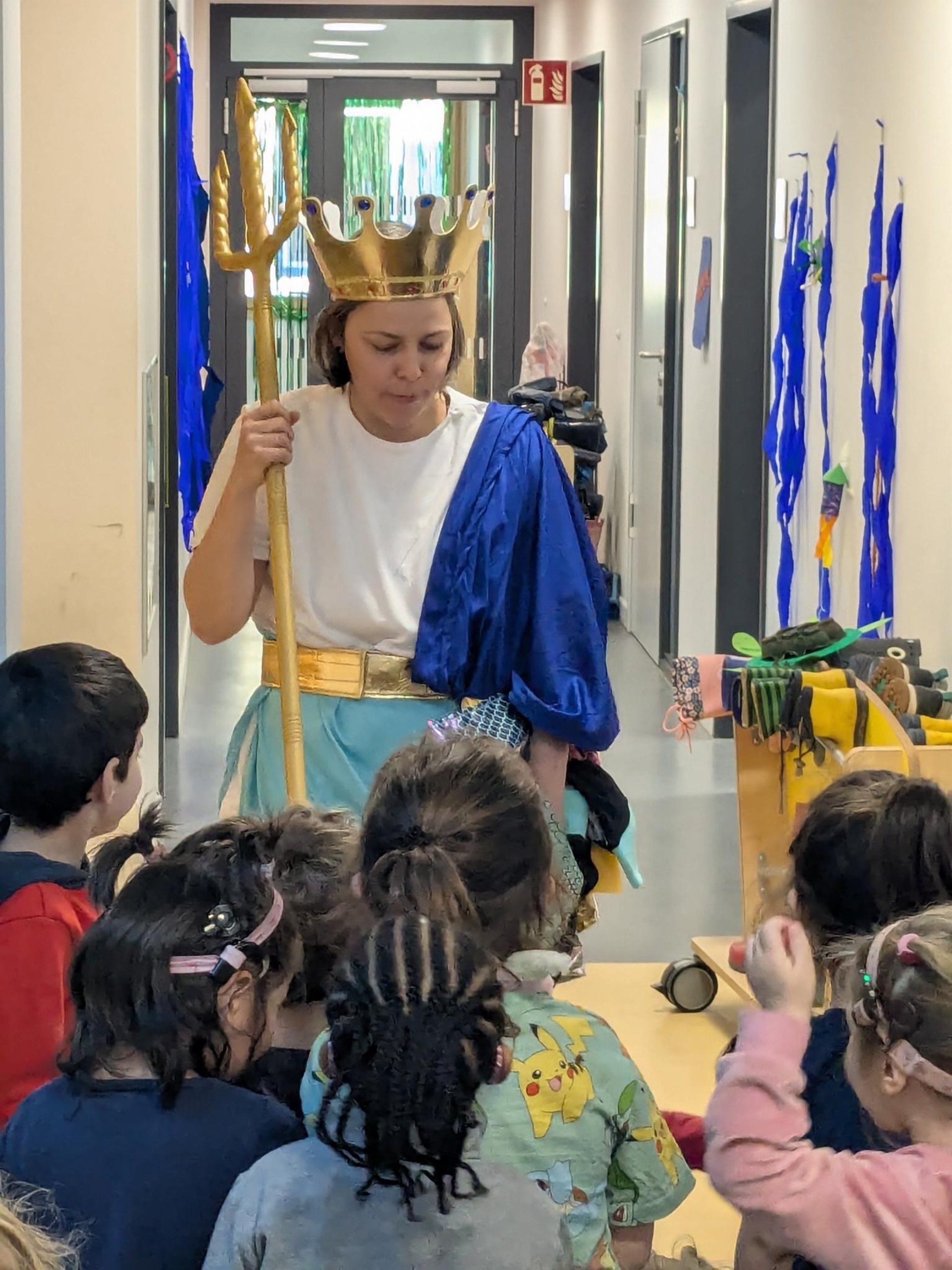 Eine Gruppe kleiner Kinder von hinten. Sie schaut zu einer Frau, die als Neptun verkleidet ist mit goldenem Dreizack und goldener Krone.