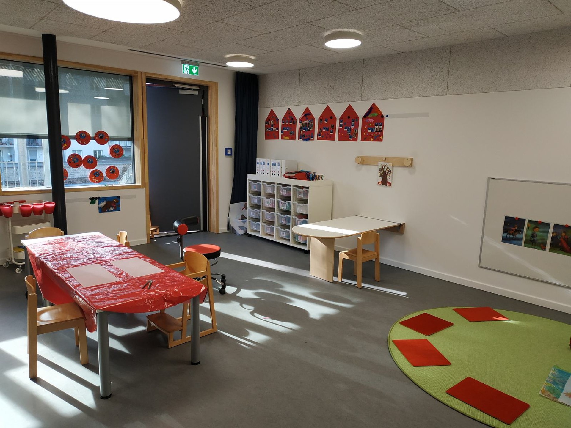 Blick in einen Raum des Kindergartens: ein Tisch mit roter Plastikdecke und Malblättern, ein runder Teppich mit Sitzkissen, Regale mit Spiel- und Bastelmaterial. An der Wand hängt ein Geburtstagskalender aus roten Häuschen.