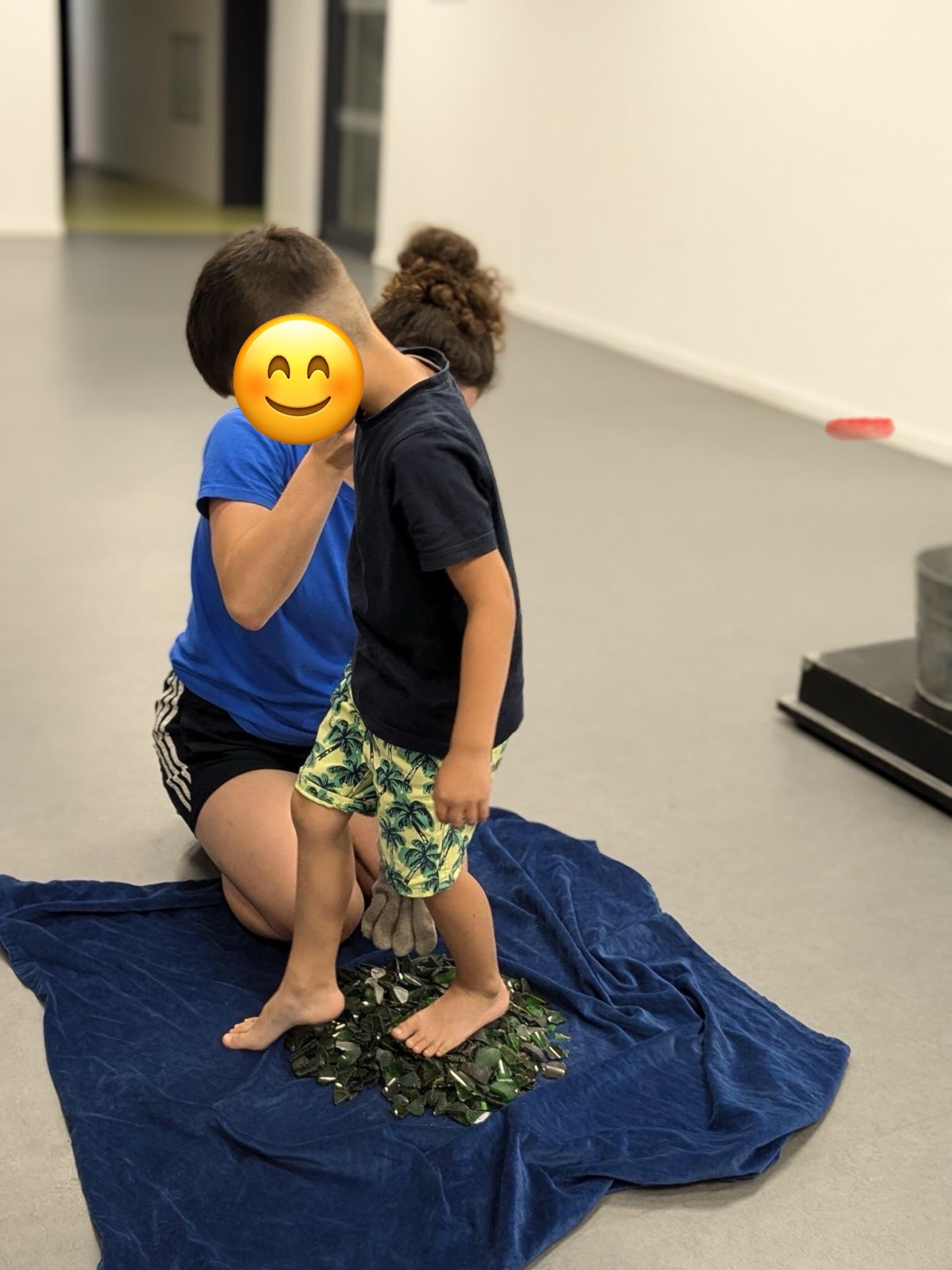 Ein Kind läuft über einen kleiner Haufen grüner Scherben, die auf einer blauen Samtdecke liegen. Eine erwachsene Person kniet neben ihm und hält seine Hand.