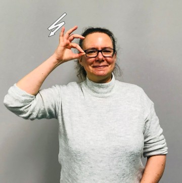 Porträtfoto einer Frau mittleren Alters mit heller Haut, brauner Krause und Brille. Gebärdenname ist FRAU EULE