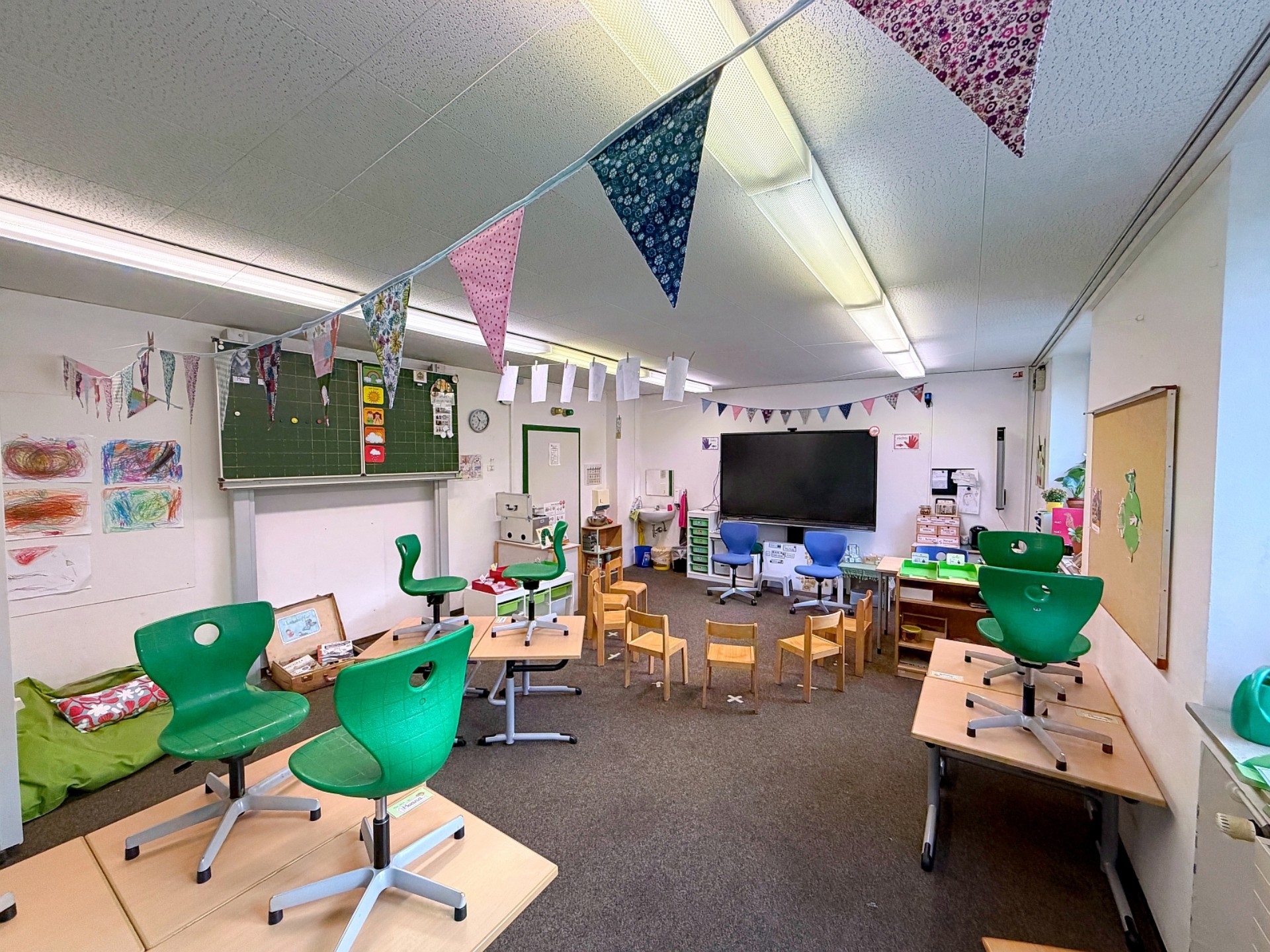 Blick in ein Klassenzimmer der Primarstufe. Bunte Stühle stehen auf den Tischen, an der Decke hängen Wimpelketten. An den Wänden sind viele Plakate und Tafeln.