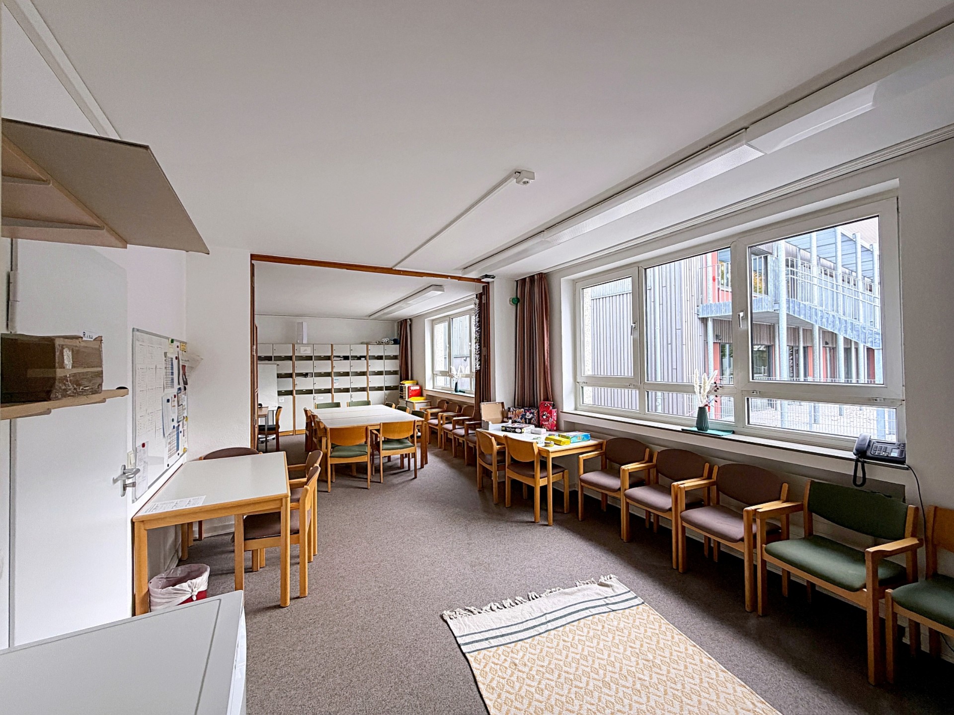 Blick in das Zimmer der Lehrkräfte: Es gibt mehrere Gruppentische, an der Wand vor den Fenstern stehen aufgereiht gepolsterte Stühle.