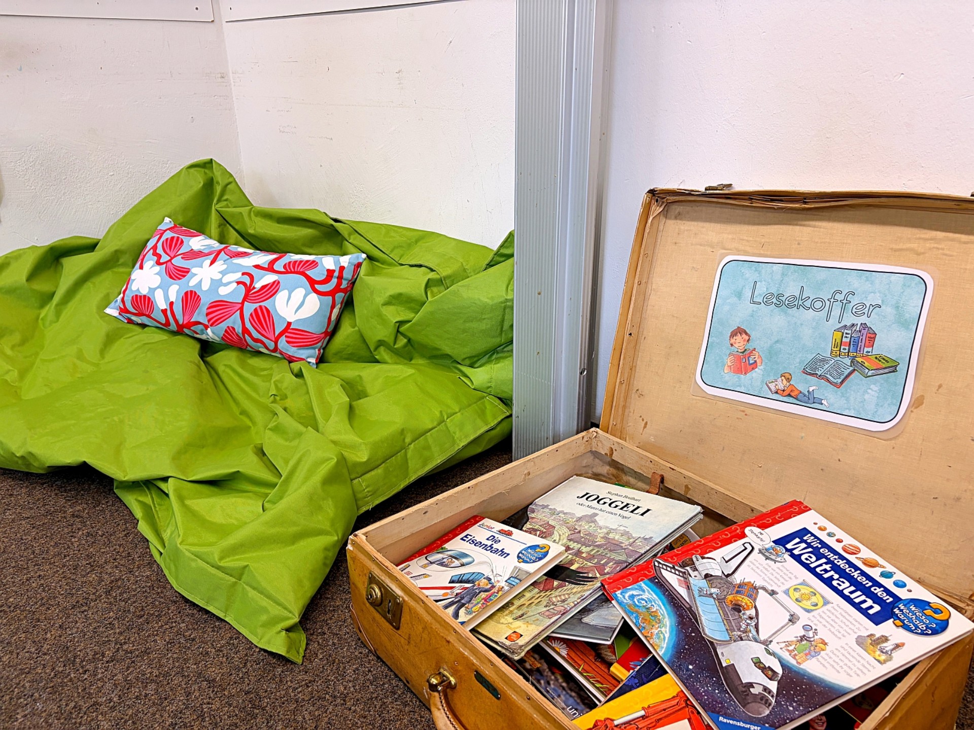 Ein großes grünes Sitzkissen und ein aufgeklappter "Lesekoffer" mit vielen Büchern