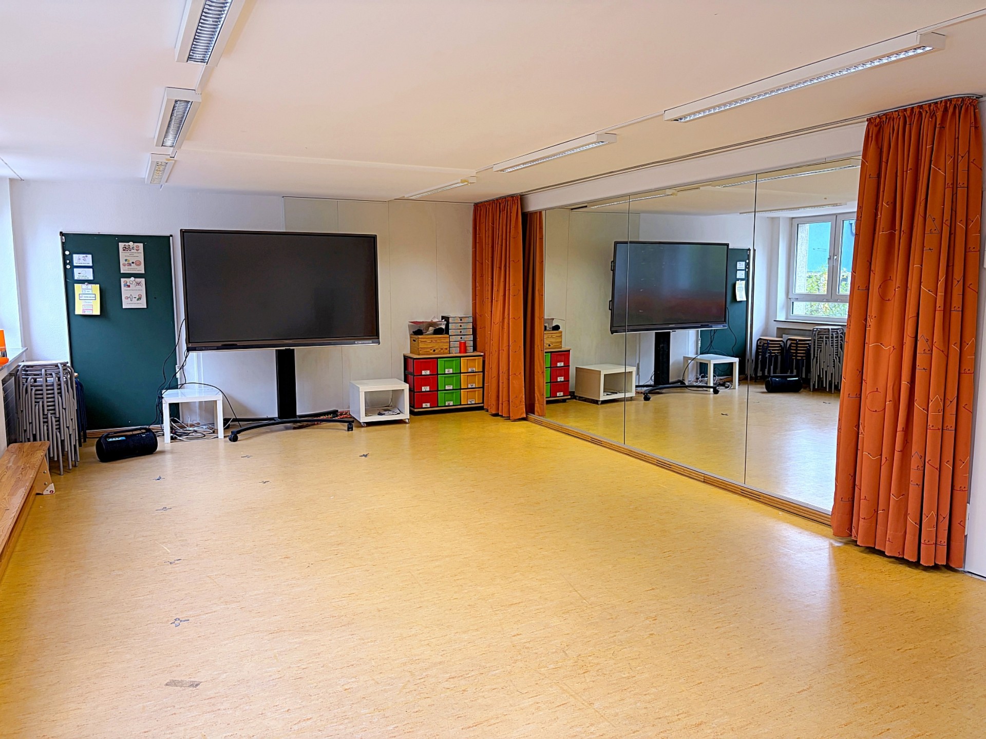 Ein großer Raum mit freigeräumten Fußboden, Hocker sind in der Ecke gestapelt. Die rechte Wand ist eine Spiegelwand, die mit einem Vorhang verdeckt werden kann.