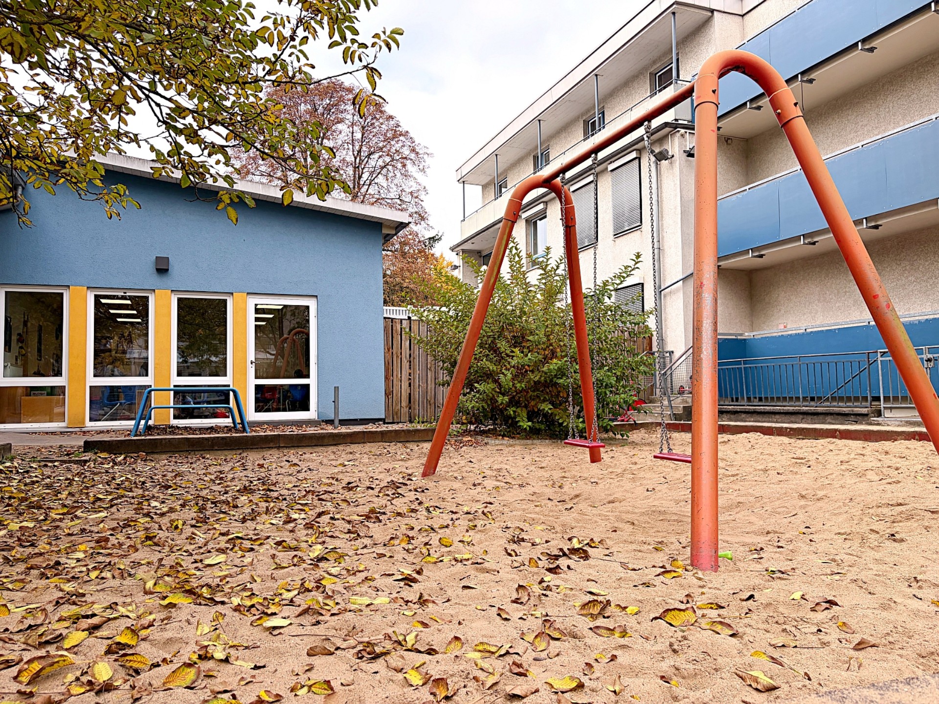 Eine rote Schaukel auf einer Sandfläche. Im Hintergrund ein Spielgerät und das blaue Haus der OGS.