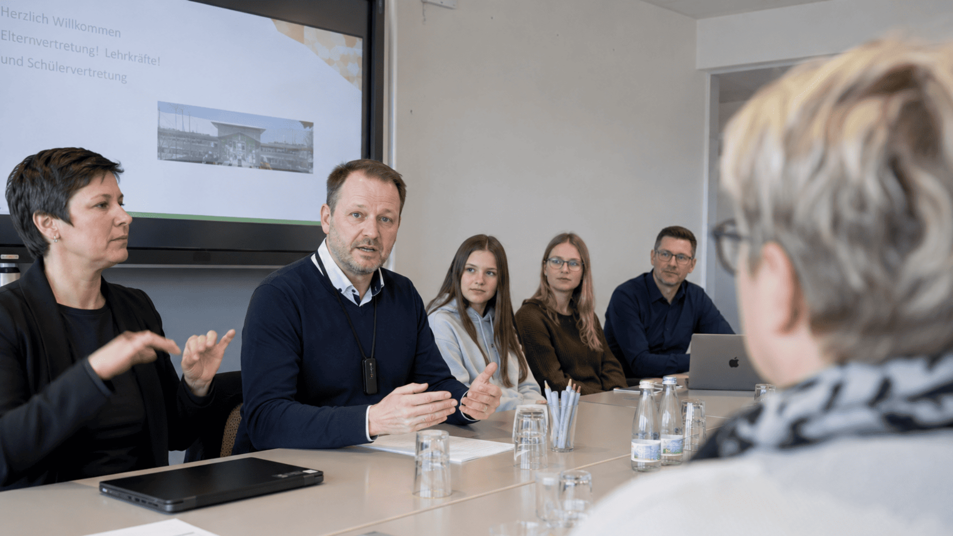 Teilnehmende einer schulischen Konferenz sitzen gemeinsam an einem Tisch. Ein Lehrer mit Mikrofon spricht, daneben unterstützt ein DGS-Dolmetscher die Kommunikation. Eltern-, Lehrkräfte- und Schülervertretung tauschen sich im Konferenzraum aus.