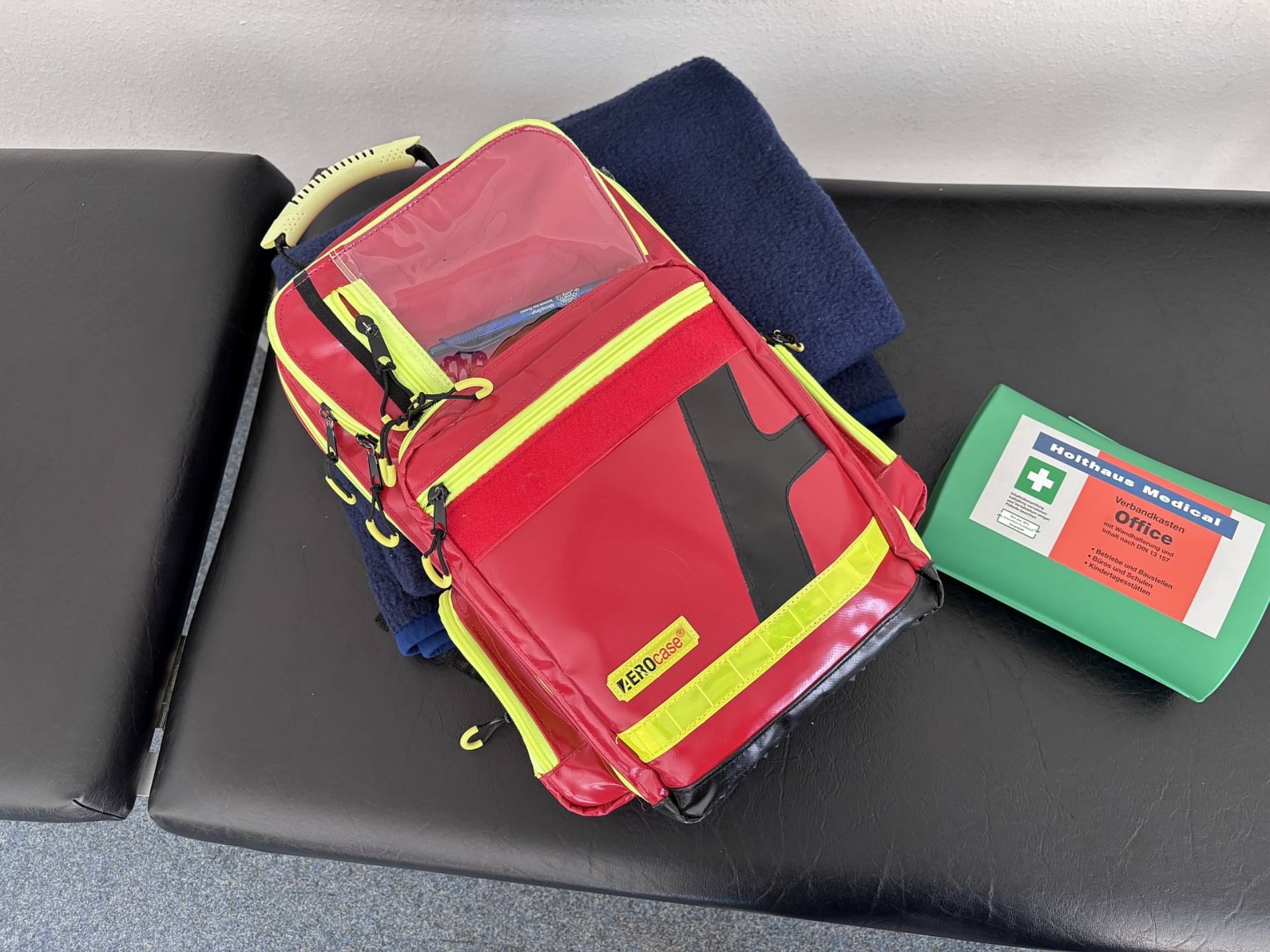 Auf einer Liege liegen ein Notfallrucksack und ein Erste-Hilfe-Päckchen.