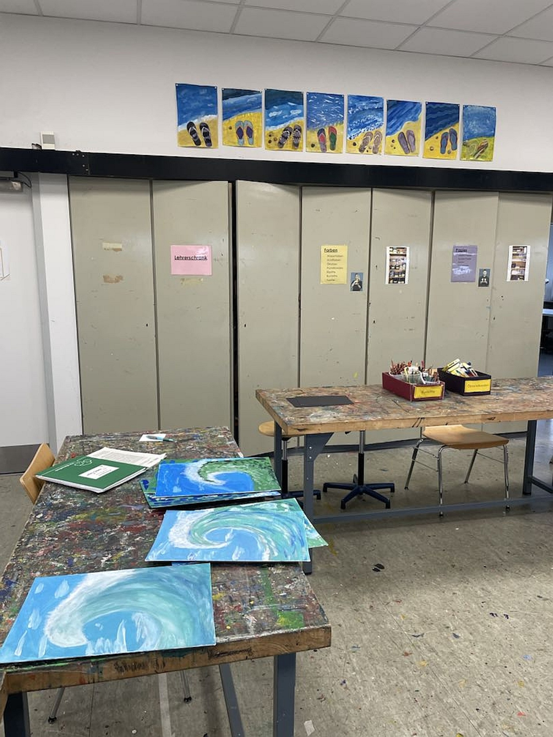 Blick in den Kunstraum: Auf den Tischen liegen Blaue Bilder von Meereswellen und Malutensilien. Im Hintergrund stehen große graue Metallschränke an der Wand.