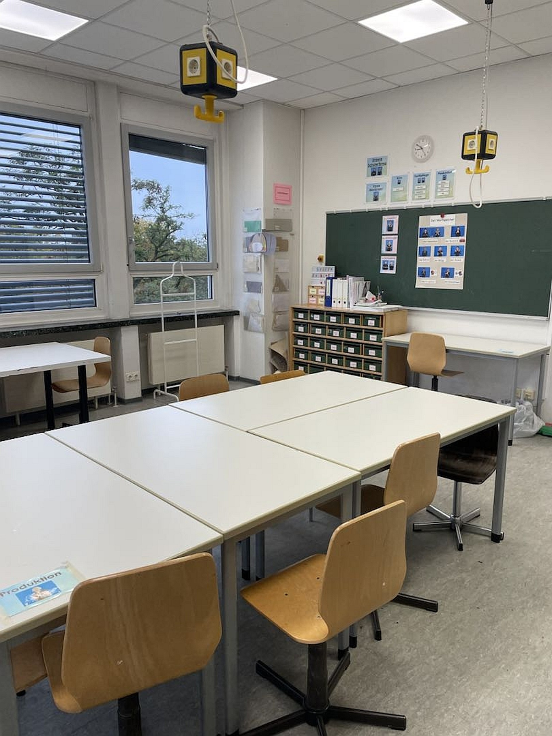 Blick in den Testilraum: Die Tische sind in der Mitte des Raumes zu einer großen Arbeitsfläche zusammengeschoben. An der Wand hängt eine grüne Tafel, darunter steht ein Regal mit Material.