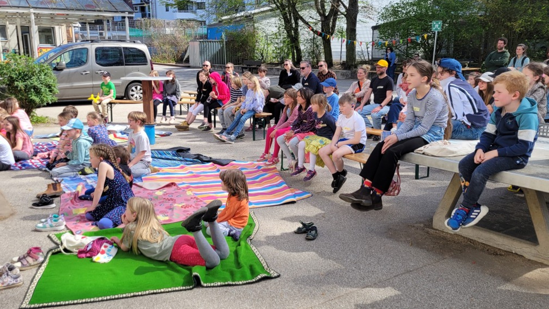 Etwa 50 sommerlich gekleidete Kinder und Eltern sitzen auf Decken, Bänken und einer Tischtennisplatte und scheun interessiert auf etwas, das nicht im Bild zu sehen ist