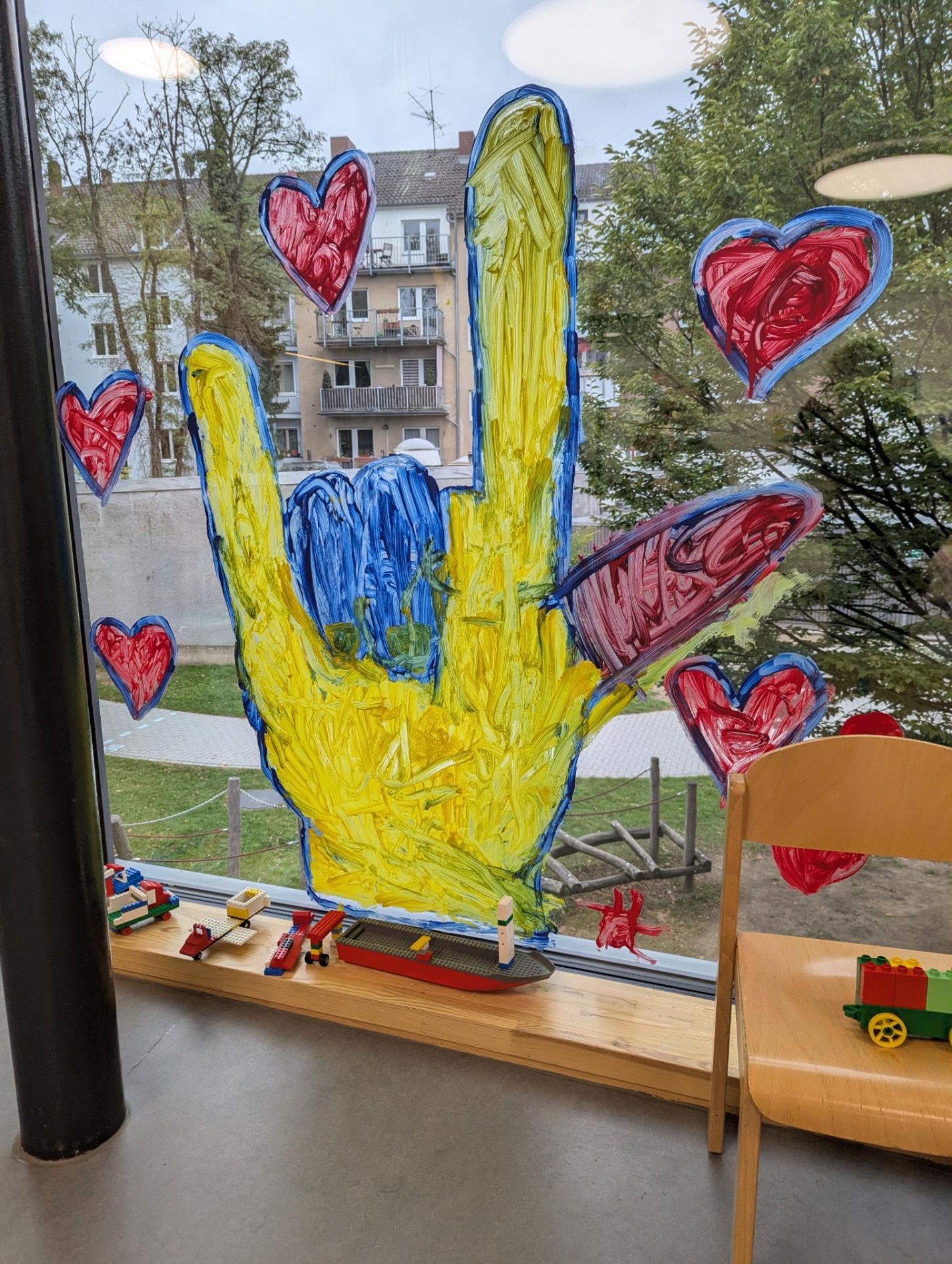 Eine  etwa 1,5 Meter hohe bunte ILY-Hand ist an ein Fenster gemalt. Um die Hand herum sind Herzen gemalt.