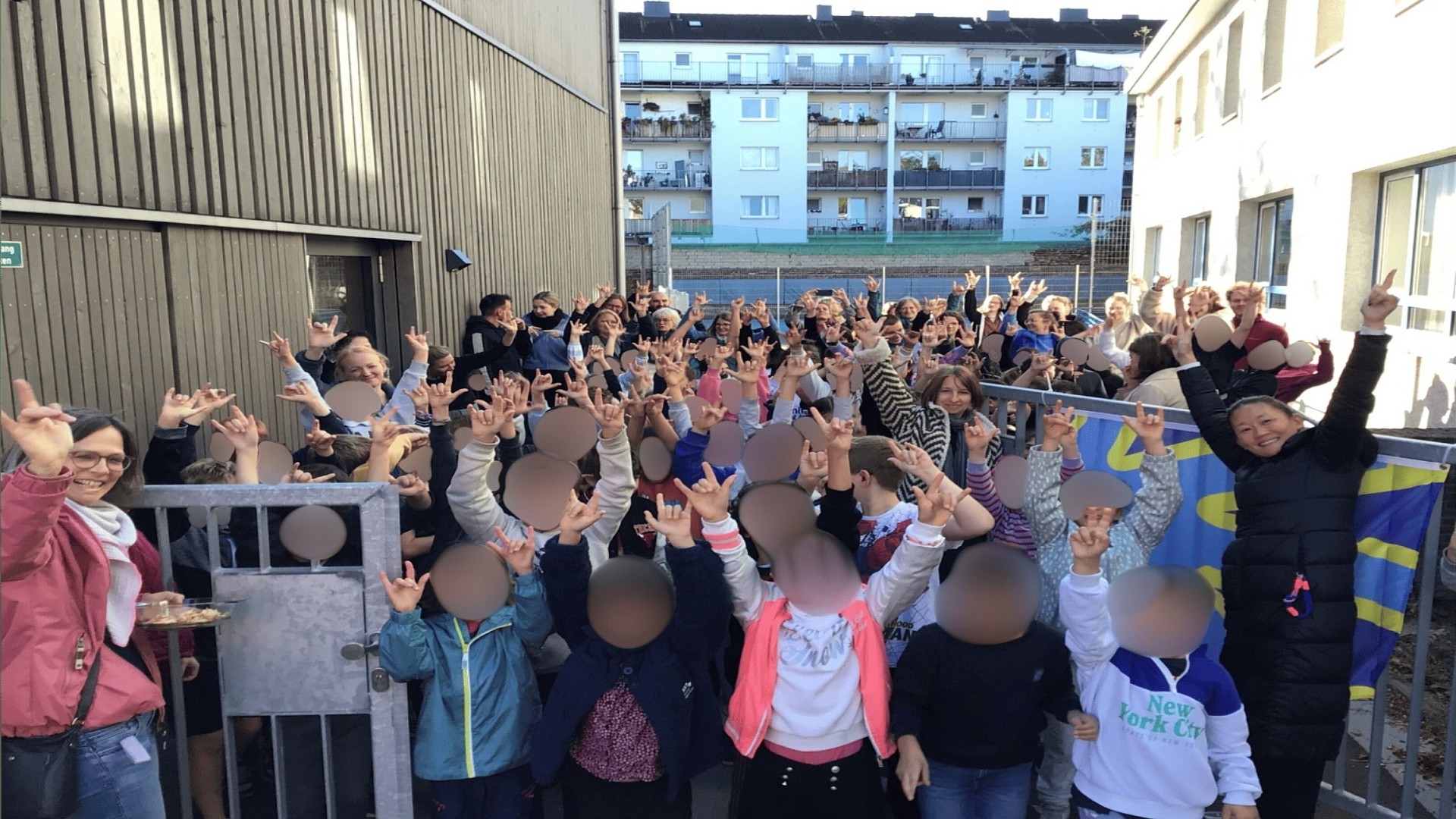 Blick auf eine große Gruppe von Kindern und Erwachsenen, die sich am Schultor zwischen zwei Gebäuden drängt: alle strecken die Hände mit der ILY-Gebärde in die Luft..