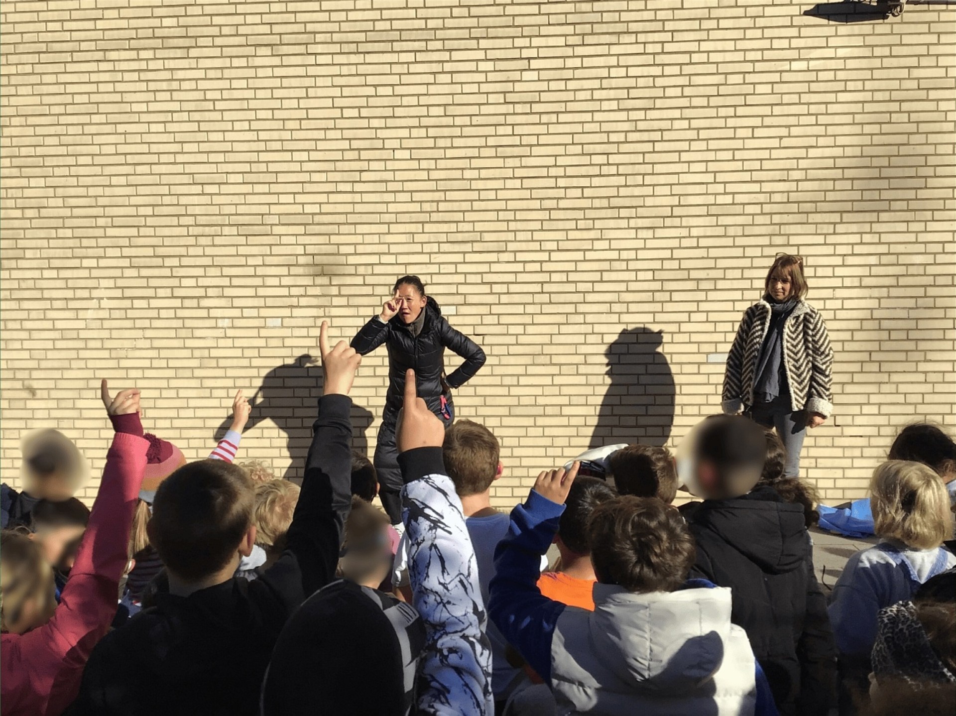 Die Schulleiterin steht vor einer Gruppe von Kindern und stellt mit einer Handgebärde eine Frage.