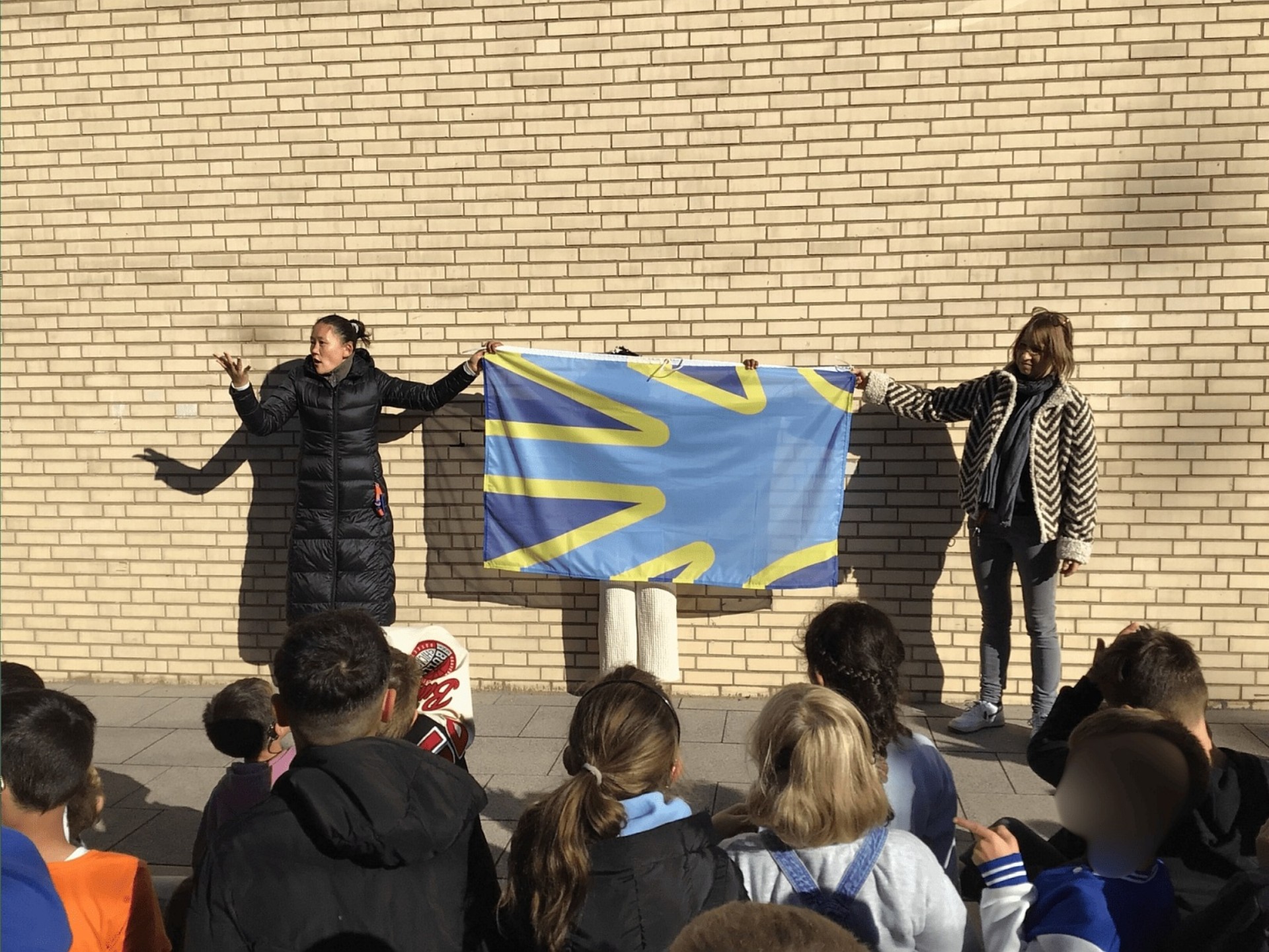 Zwei Frauen stehen vor einer verklinkerten Wand. Sie halten die blaue Gehörlosen-Flagge. Vor ihnen stehen viele Kinder, die man nur von hinten sieht.