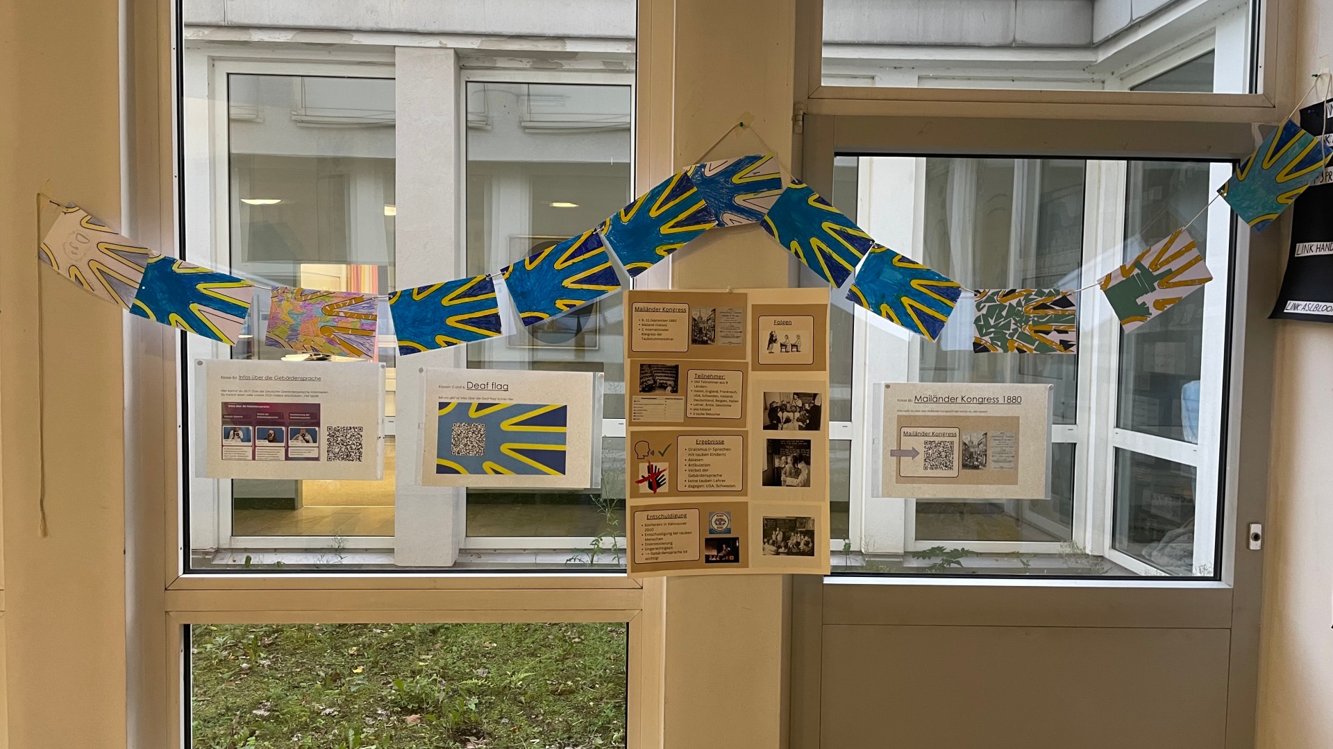 Vor einem Fenster hängen DIN A6-große Wimpel mit bunt gestalteten Gehörlosen-Flaggen und Infoplakate zu dieser Flagge.